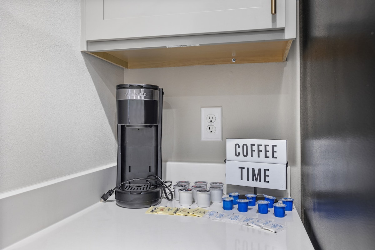 A coffee station is presented with a black coffee maker placed on a white surface. Various coffee pods are neatly organized nearby, alongside a selection of sugar packets. A lightbox displays the text 'COFFEE TIME', contributing to a functional beverage area.