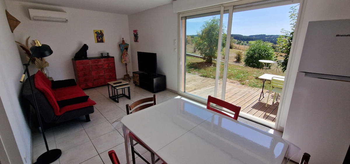 A bright living area features large glass doors opening to a scenic view. A red sofa is positioned beside a sleek black TV stand, while a dining table with red chairs is visible. Light-colored tiles add to the spacious feel.