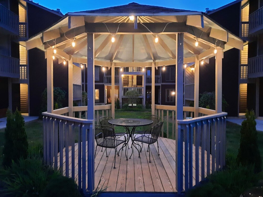 A wooden gazebo is illuminated by soft string lights, featuring a round table and four chairs on the deck. The surrounding greenery adds a natural touch, while the two-story buildings create a cozy courtyard ambiance.
