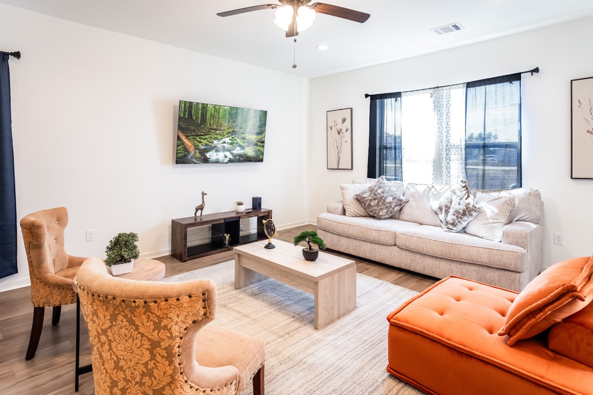 A spacious living room features a light-colored couch and an orange lounge chair. Two upholstered chairs are positioned beside a wooden coffee table. A large television, displaying a nature scene, is mounted on the wall, with natural light filtering through the windows adorned with sheer curtains.