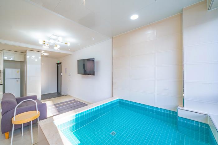An indoor area features a compact pool with clear turquoise water, bordered by tiled flooring. A light gray sofa sits adjacent to the pool. A flat-screen television is mounted on a white wall, and natural light is streamed through the space, enhancing its clean and modern design.