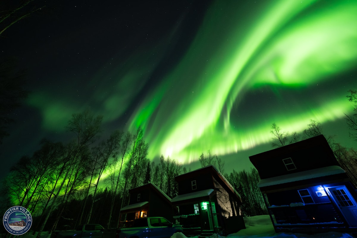 The image captures vibrant green auroras swirling across a night sky, illuminating the landscape. Two guest cabins, partially covered in snow, are visible in the foreground, with soft lighting emanating from their windows. Surrounding birch trees contribute to the serene atmosphere of the rural setting.