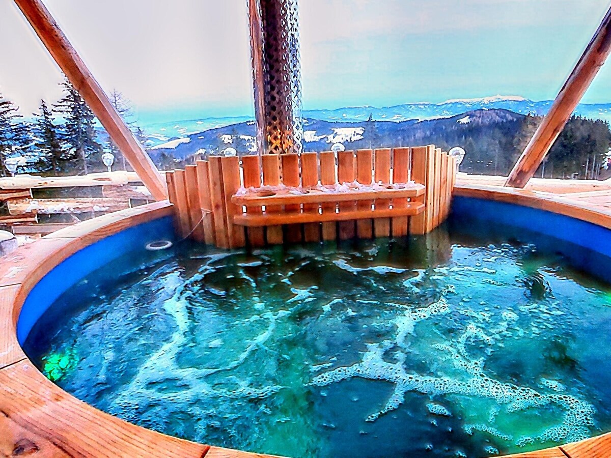A wooden hot tub is presented with bubbling water, surrounded by a scenic view of mountains and valleys. The open structure features wooden beams and seating, allowing for an immersive outdoor experience in a serene alpine setting.