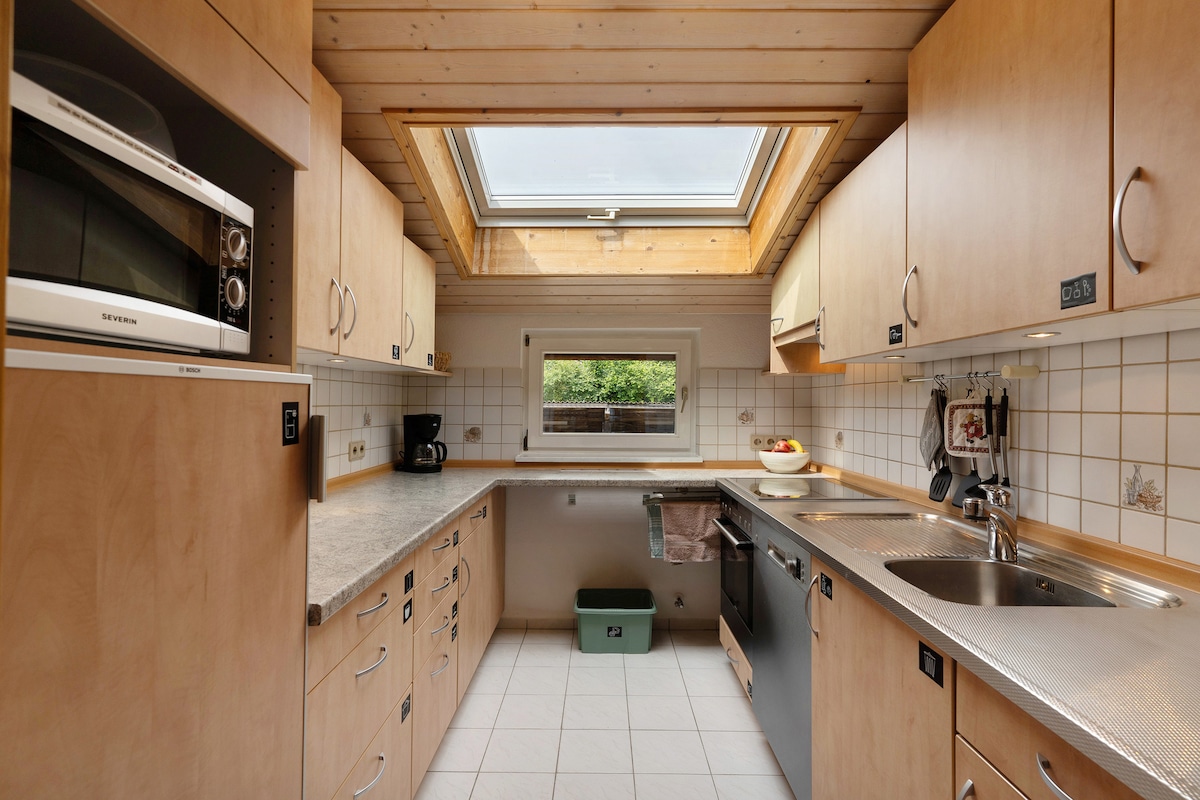 The kitchen is designed with light wood cabinetry and a spacious countertop. A skylight allows natural light to enhance the workspace. Key appliances include a microwave, coffee maker, and dishwasher, with a window providing a view of the outdoor greenery.