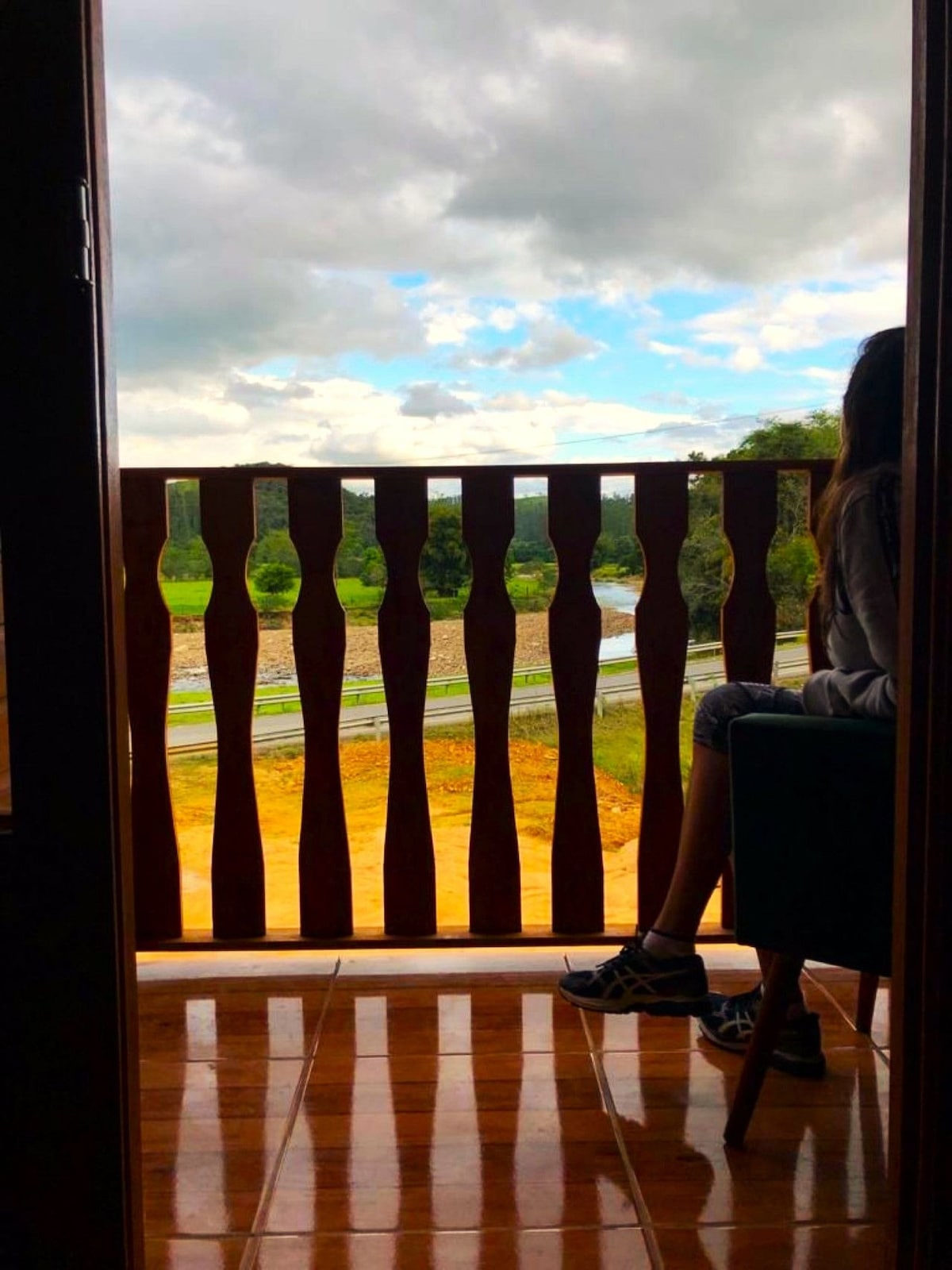A balcony with wooden railing offers views of the surrounding landscape. The scene features green fields and a river, with clouds partially covering the sky. A person is seated, partially visible, enjoying the serene atmosphere of the outdoors.