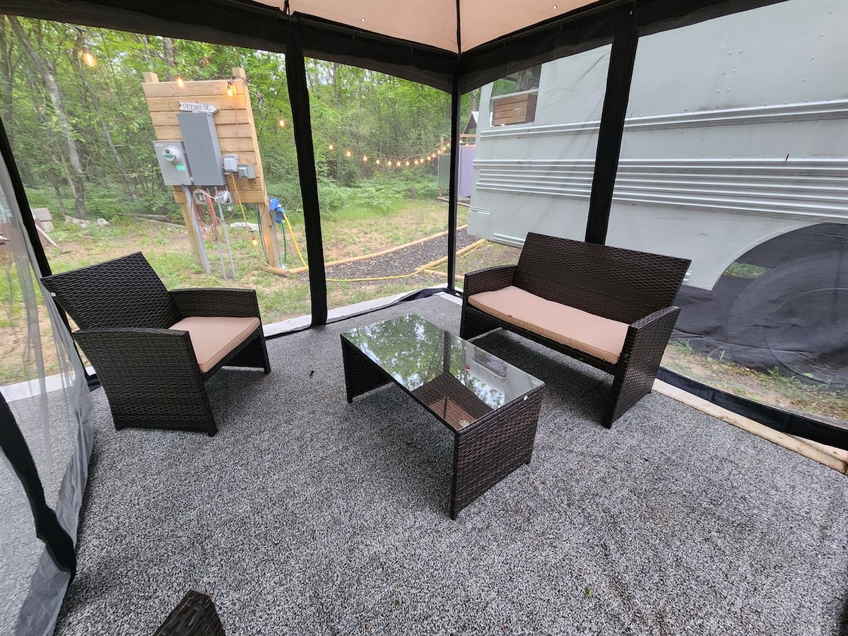 A screened outdoor space features two wicker chairs with neutral cushions and a low glass table at the center. The floor is covered in textured gray carpet. Soft outdoor lighting can be seen in the background, enhancing the area’s connection to nature.