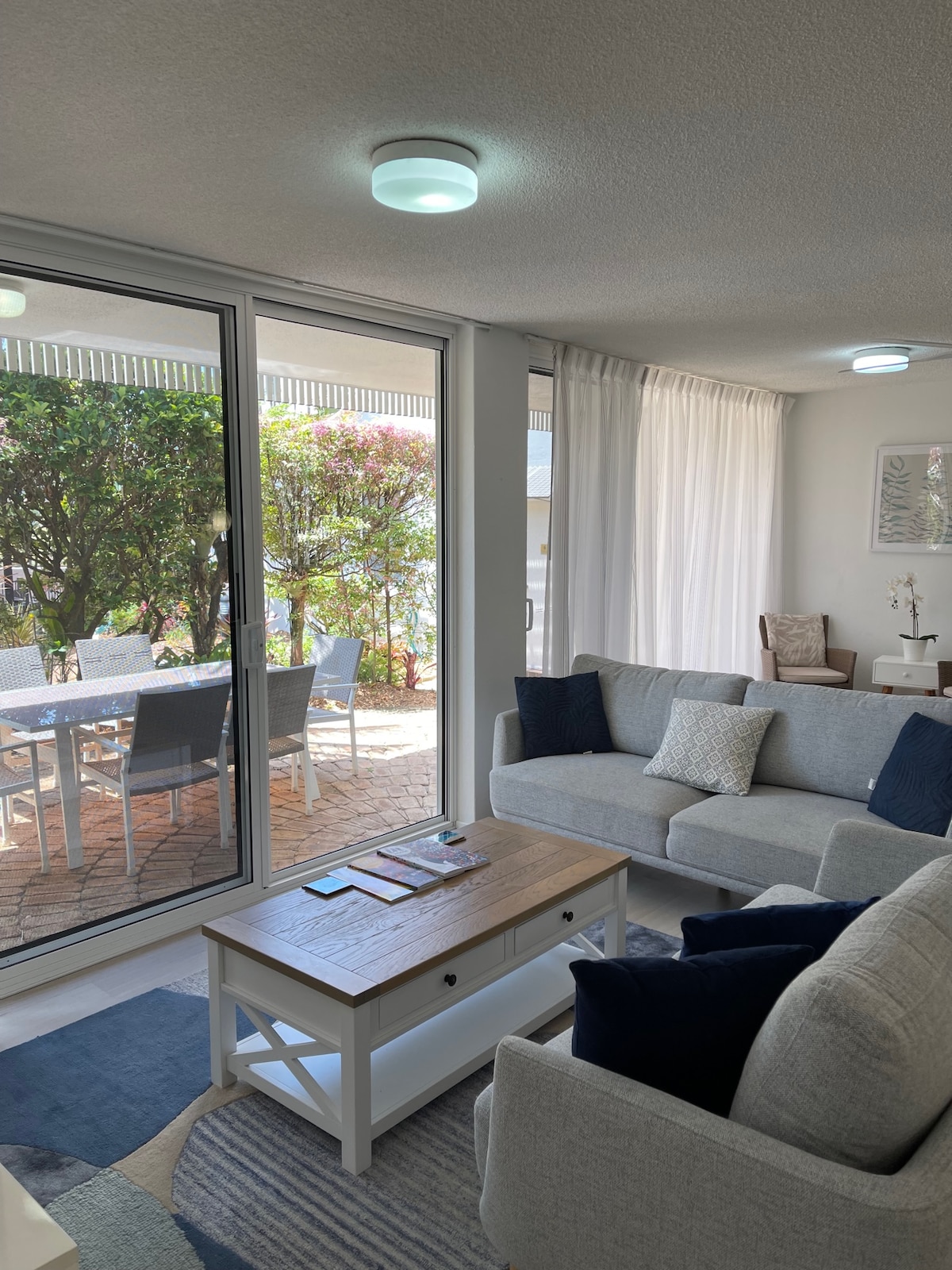 A living area features a light grey sofa paired with navy accent pillows, complemented by a wooden coffee table. Large glass doors provide a view of a private patio with outdoor dining, framed by lush greenery and soft natural light.