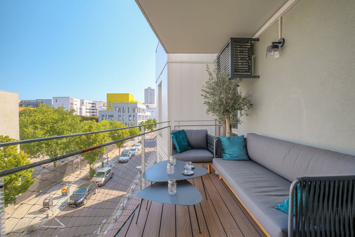 An elegant balcony space is showcased, featuring a comfortable grey sofa adorned with teal cushions. A glass table holds decorative candles, while surrounding trees provide a touch of greenery. The view includes nearby buildings and a quiet street below.