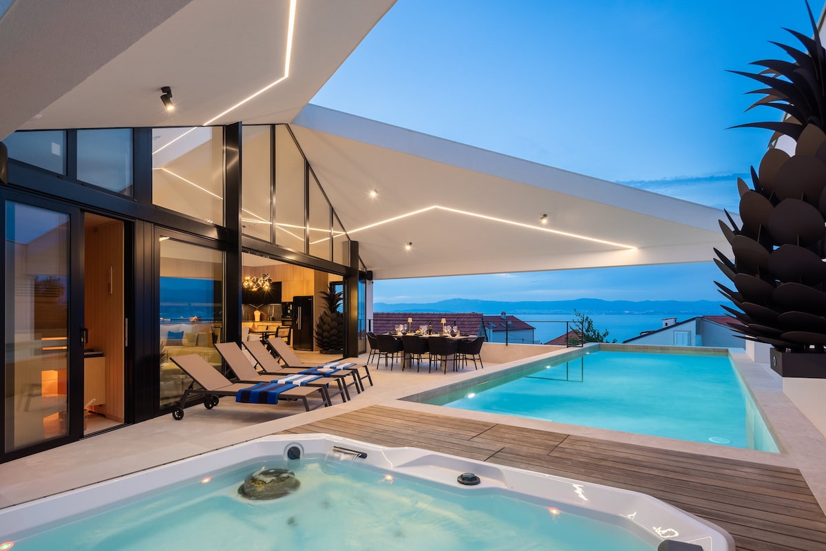 An outdoor area showcases a spacious private pool and an adjacent outdoor jacuzzi, illuminated by soft lighting. Lounge chairs are placed near the pool, and a dining table for eight is positioned under a sleek overhanging roof, providing comfort and a view of the sea.