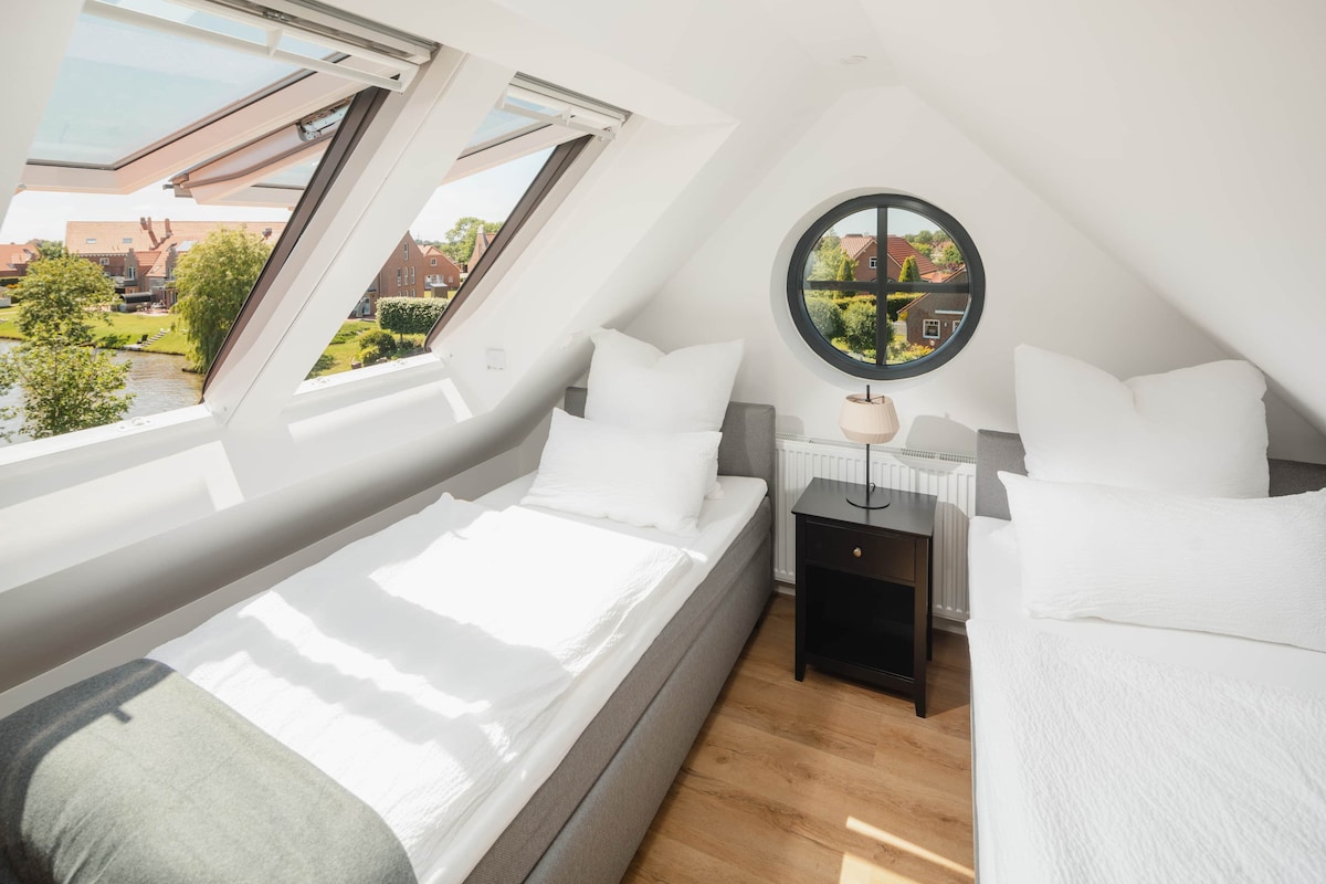 A well-lit attic room features two separate beds with white bedding positioned beside a round window. Natural light floods the space through multiple skylights, highlighting the light-colored wooden flooring and a bedside table with a lamp.