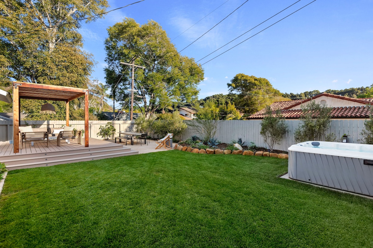 A spacious backyard features a well-maintained green lawn, surrounded by shrubs and trees. A wooden deck with seating is present to one side, while a hot tub is positioned on the opposite side, offering a relaxing outdoor space.