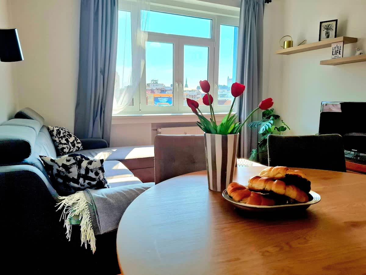 A bright living space is equipped with a comfortable sofa and a wooden dining table. A vase with red tulips decorates the table, alongside a plate of pastries. Large windows allow natural light to illuminate the room, enhancing the view of the city beyond.
