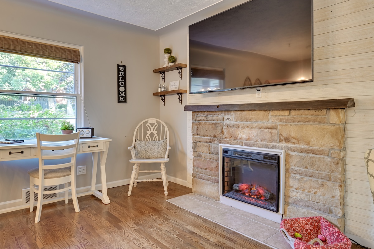 A cozy living area is highlighted by a modern electric fireplace embedded in a stone wall. A large flat-screen TV is mounted above the fireplace. A small desk is positioned near a window, with natural light illuminating the hardwood floor.