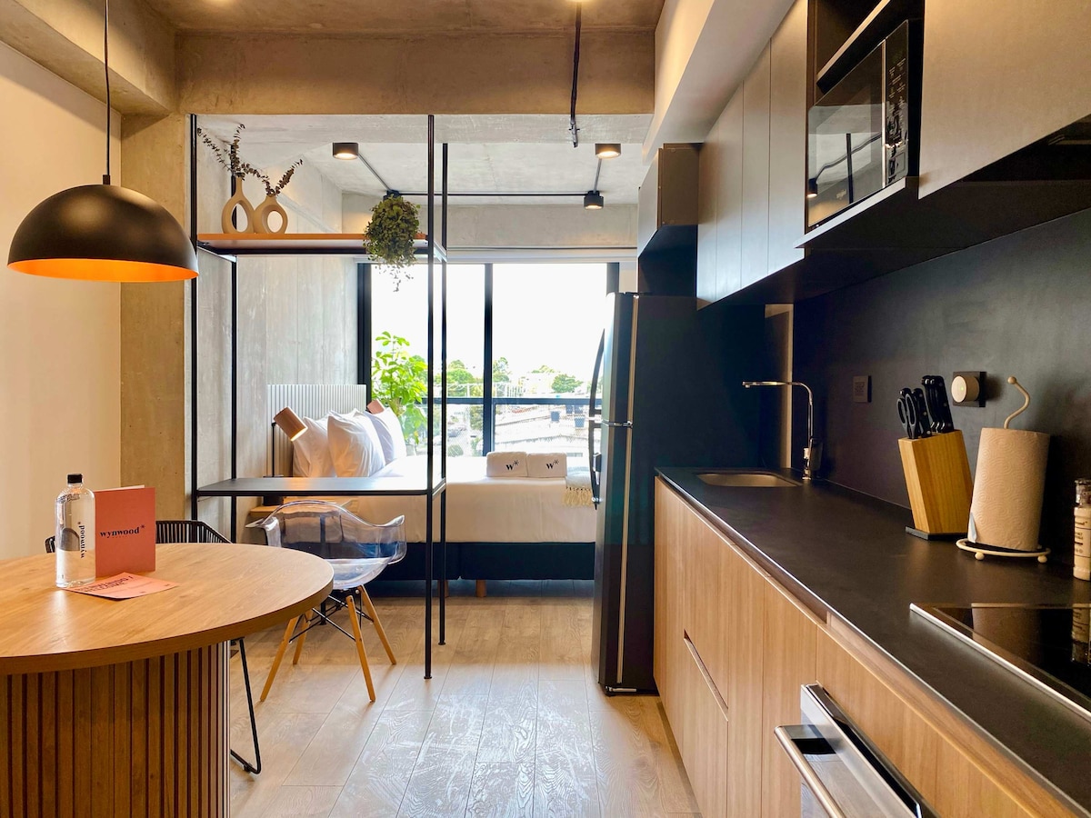 A modern kitchen area is highlighted by sleek cabinetry and essential appliances. A round wooden dining table is set to one side, with a clear plastic chair nearby. Large glass doors lead to a bright outdoor space, revealing a cozy sleeping area with fresh linens.