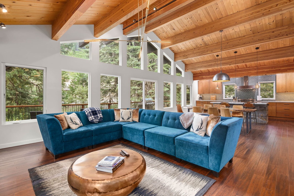 The living area features a large, plush wrap-around couch in a blue hue, complemented by decorative pillows. Natural light floods the space through multiple windows, enhancing the wood-beamed ceilings. A round coffee table sits at the center, with a view of the open-concept kitchen in the background.