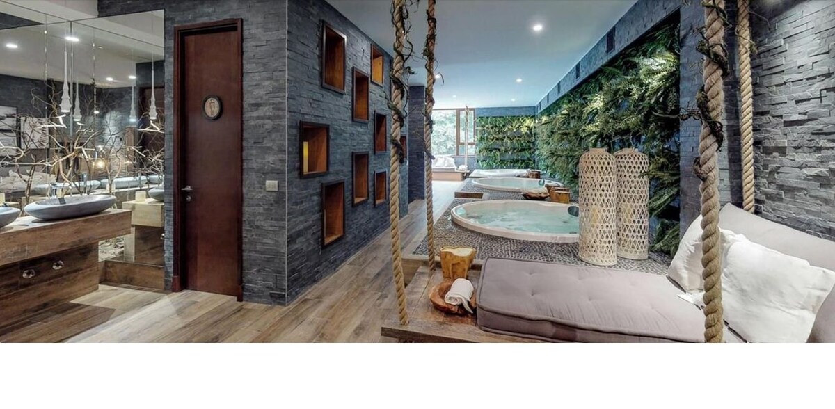 A modern wellness area is displayed, featuring elegant stone walls and natural wood accents. Two hot tubs are positioned beside lush greenery, enhancing the tranquil setting. Comfortable lounging areas with soft cushions invite relaxation, while suspended swings add a unique touch to the design.