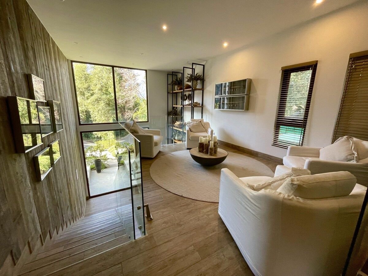 A spacious living area features large windows that frame views of the outdoor greenery. Two comfortable white armchairs are placed around a circular coffee table on a round area rug, enhancing the airy ambiance. Natural light fills the space, highlighting the wooden accents.
