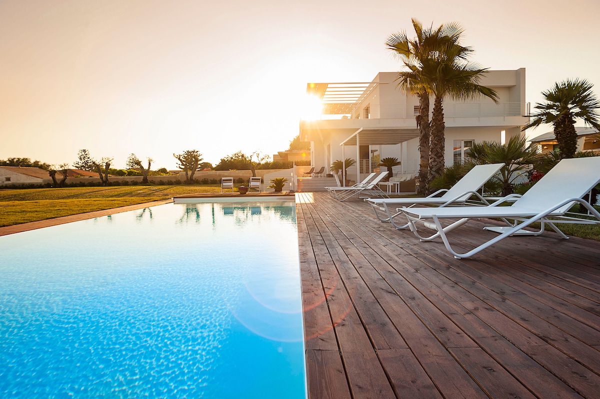 A serene view of the swimming pool is highlighted by the gentle reflection of the setting sun. Lounge chairs are positioned on a wooden deck, surrounded by landscaped greenery, creating a relaxing outdoor environment.