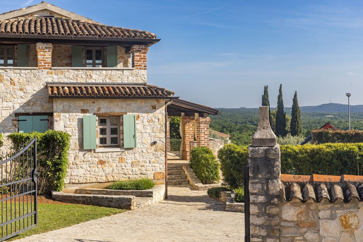 The exterior of the villa showcases a blend of traditional stone and brickwork, accented by green shutters. A well-maintained pathway leads to the entrance, framed by lush greenery and cypress trees. The surrounding landscape features rolling hills under a clear blue sky.