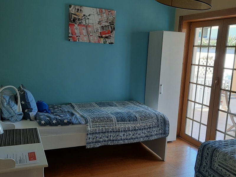 A bright room features two twin beds with patterned bedding. A white wardrobe stands beside the beds. A large window with multiple panes opens to a terrace area. A piece of wall art adds a pop of color against the blue wall.