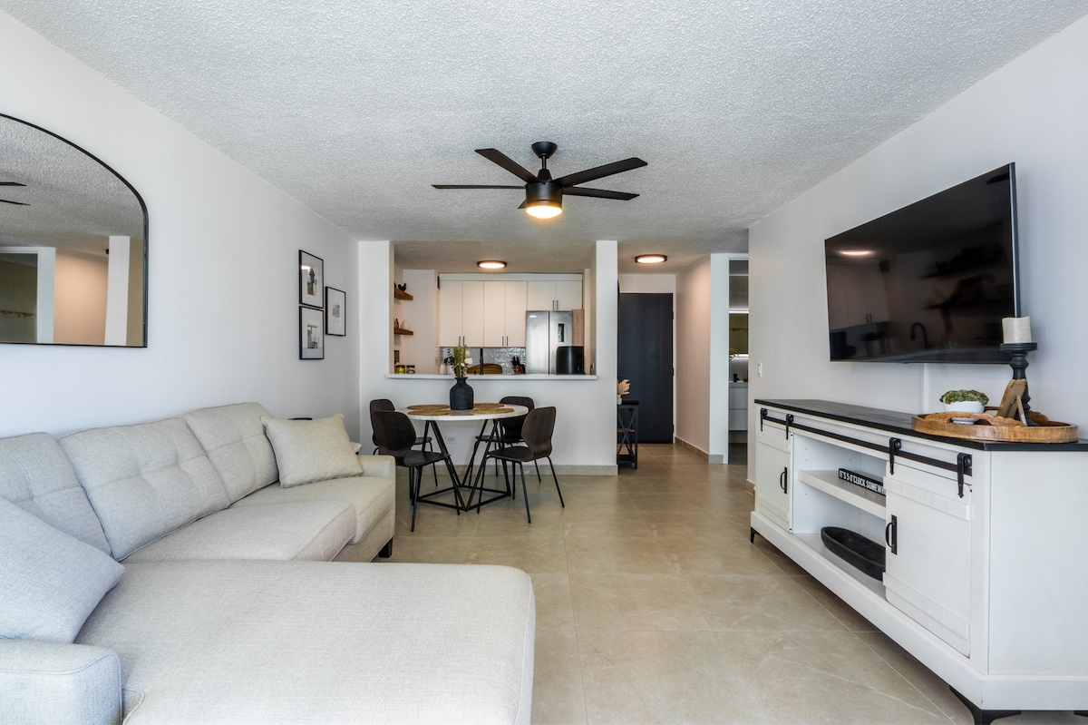 A spacious living room features a light-colored sectional sofa and a round dining table surrounded by four chairs. A large flat-screen TV is mounted on a white entertainment center. The open kitchen layout is visible in the background, allowing natural light to fill the space.