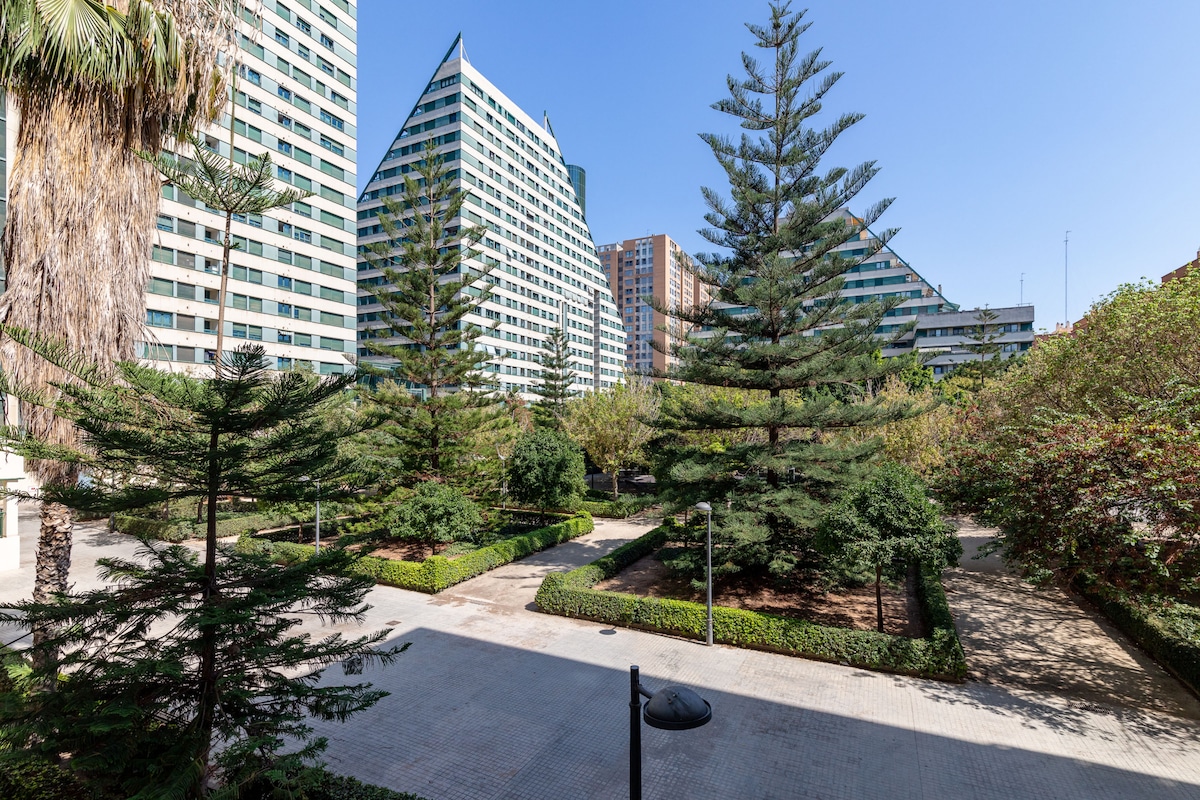 The image displays a serene garden square framed by lush greenery and tall palm trees. Modern buildings rise in the background, providing a contemporary contrast to the natural landscape. The scene reflects an inviting outdoor space, ideal for relaxation and unwinding.
