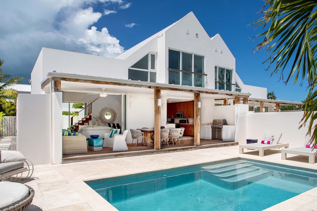 The outdoor living area is highlighted by a private saltwater swimming pool, surrounded by lounge chairs and enveloped by a tropical garden. The villa features large glass windows, allowing natural light to fill the interior space, which includes a dining area and a modern kitchen.