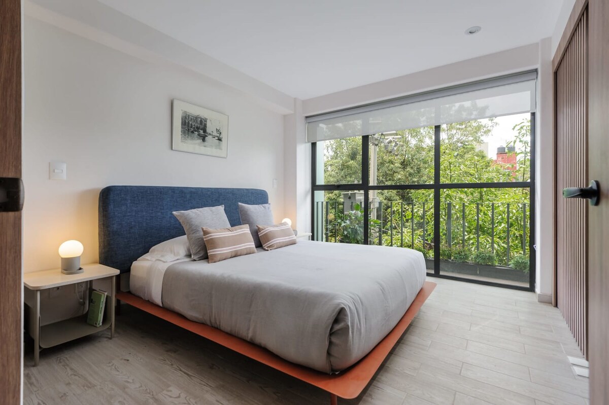 A cozy bedroom features a spacious bed with a blue upholstered headboard and soft linens. Natural light streams through large glass doors that provide a view of surrounding greenery. A small nightstand with a lamp is positioned beside the bed, enhancing the serene atmosphere.