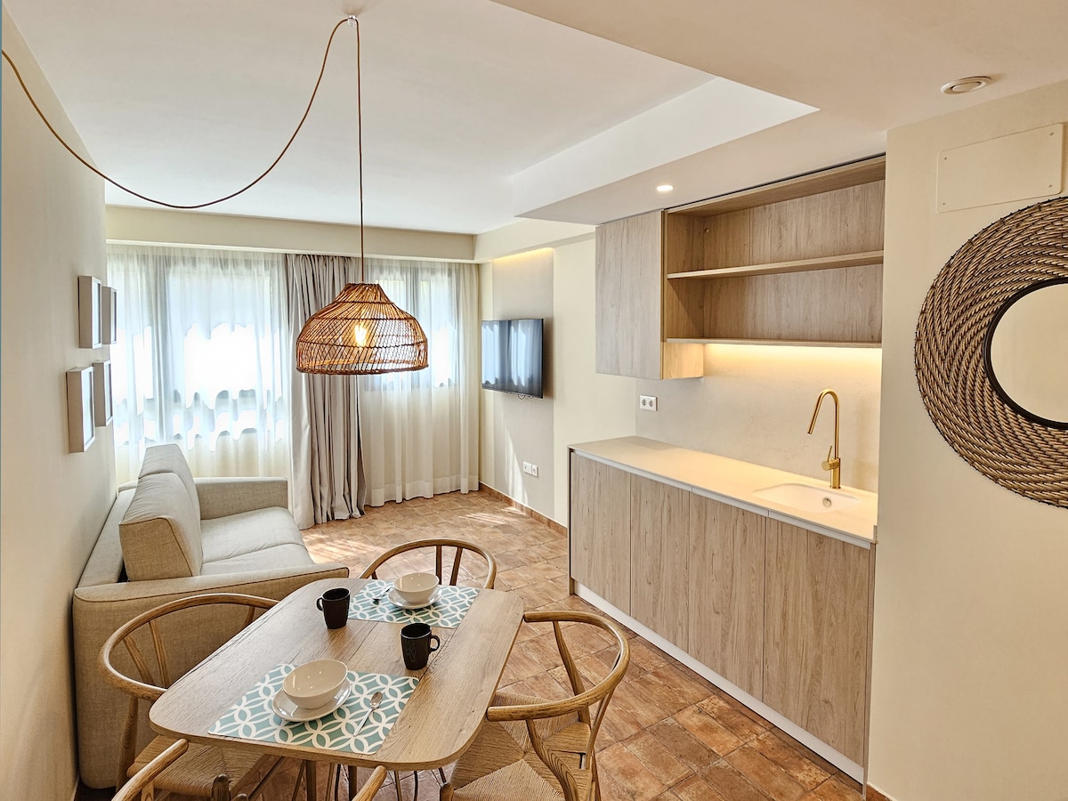 A bright and modern living-dining area features a comfortable sofa and a dining table set for four. Natural light filters through sheer curtains, highlighting warm tones in the decor. A well-appointed kitchen extends along one wall with neutral cabinetry, enhancing the spacious feel.