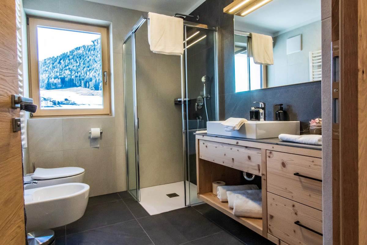 The modern bathroom features a glass shower enclosure and a stylish sink atop a wooden vanity. Natural light streams through a window, illuminating the space and providing a view of the surrounding mountains. Two bidets and neatly arranged towels add to the room's functionality.