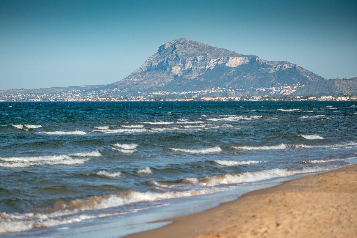 A scenic view showcases a sandy beach with gentle waves lapping at the shore. In the background, a prominent mountain rises against the clear blue sky, framed by a distant coastal city. The colors create a serene coastal landscape.