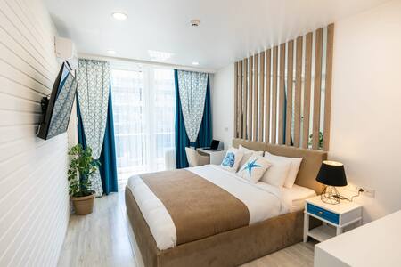 A well-lit bedroom features a queen-size bed with neutral bedding and decorative pillows. Large windows dressed with blue curtains allow natural light to fill the space. A wall-mounted television and a small desk create a functional layout, with a potted plant adding a touch of greenery.