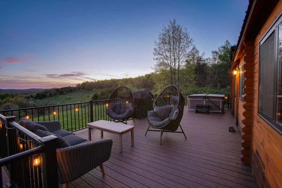 A spacious deck offers comfortable wicker seating with two hanging chairs and a rustic table. A propane grill is positioned nearby for outdoor cooking. Warm lights illuminate the area, providing a cozy ambiance against a backdrop of expansive views of the natural landscape.
