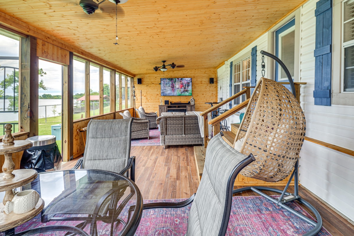 A spacious screened porch is featured with comfortable seating arrangements and a flat-screen TV mounted on the wooden wall. Large windows provide views of the surrounding nature and the waterfront, while wooden ceiling beams and fans contribute to a relaxed atmosphere.