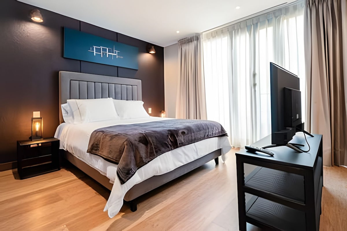 A modern bedroom features a spacious bed with crisp white linens and a dark grey throw blanket. Large windows invite natural light, contrasting with the deep blue accent wall. A TV stands on a black console beside two lamps, creating a well-lit and tranquil ambiance.