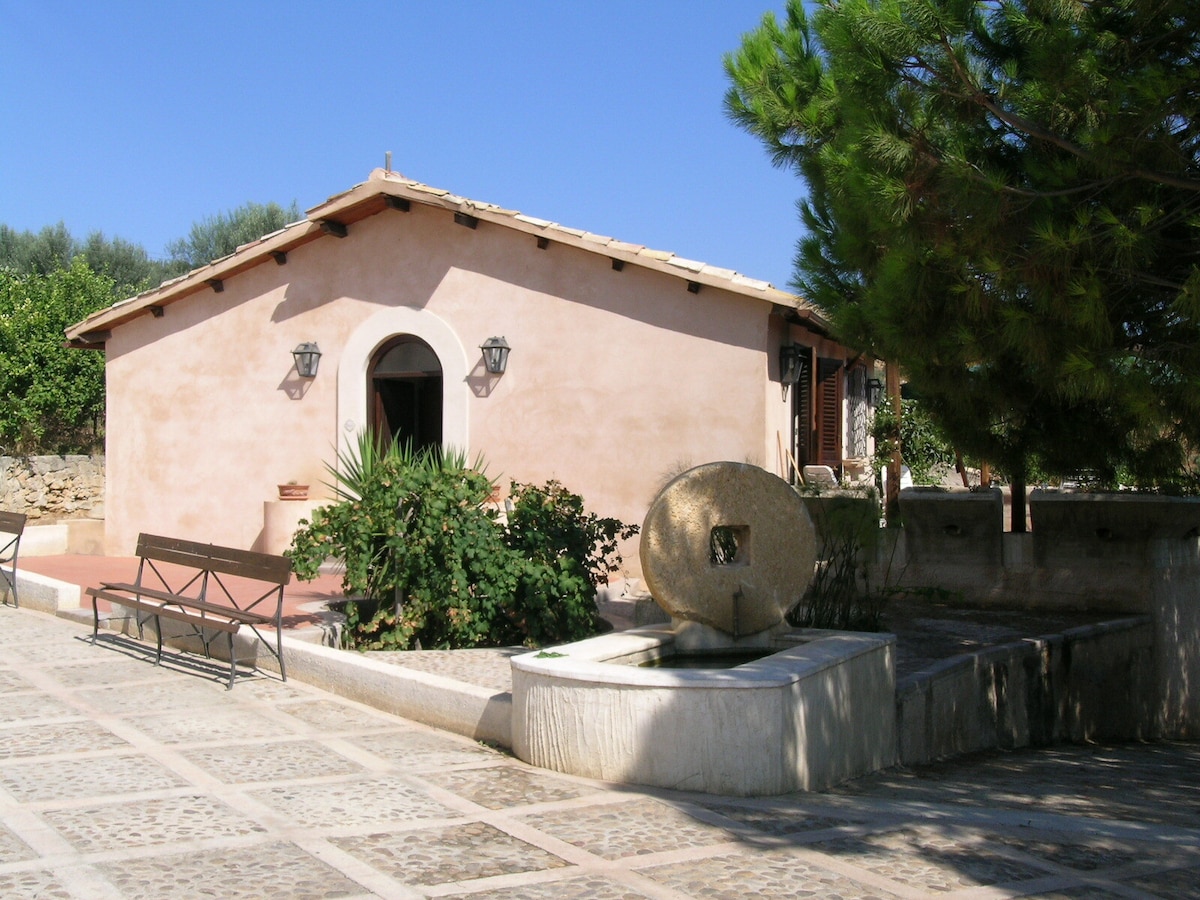 A charming farmhouse is surrounded by greenery, featuring a light-colored exterior and a terracotta roof. An inviting stone fountain is positioned near the entrance, bordered by lush plants. Benches provide seating along the pathway, which is paved with square stones.