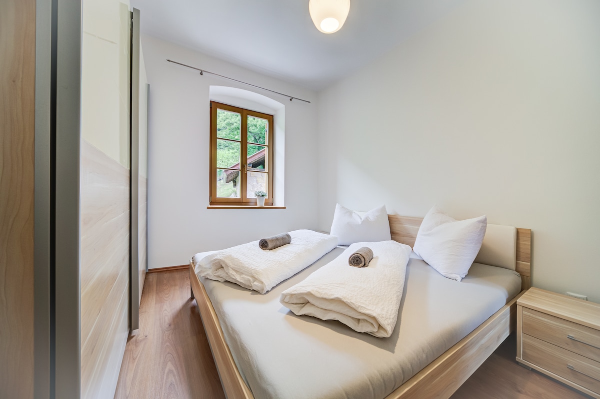 A bright bedroom is highlighted by two comfortable beds with white linens and brown towels neatly placed on top. Natural light streams in through a window with wooden framing, casting a soft glow on the light wooden furniture and warm-toned floor.
