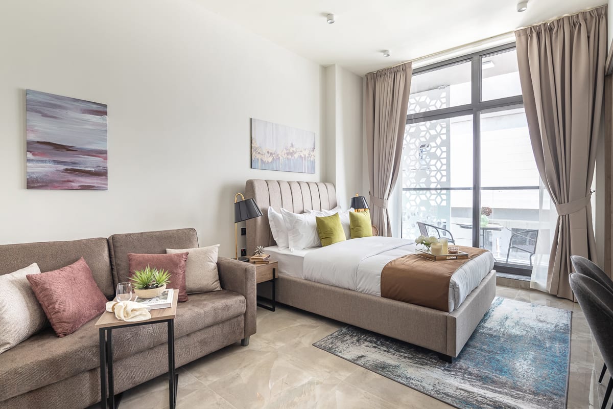 A modern bedroom is displayed, featuring a spacious bed dressed in neutral linens. A comfortable sofa is positioned beside the bed, adorned with decorative pillows. Light filters softly through large windows with sheer curtains, and a small balcony with chairs is visible in the background.