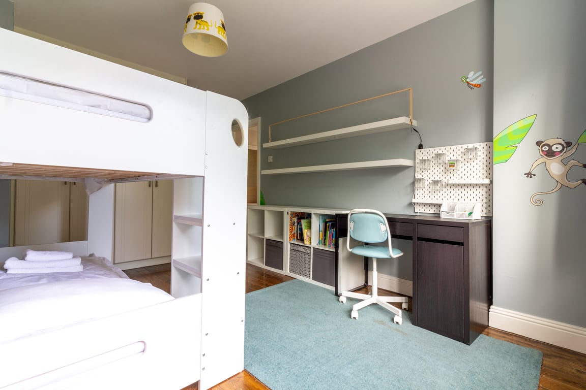A thoughtfully designed children's bedroom features a stylish bunk bed with crisp white linens. A light blue rug covers the floor, and a desk with a comfortable chair is placed next to a wall adorned with playful monkey decor. Shelving is provided for storage and organization.