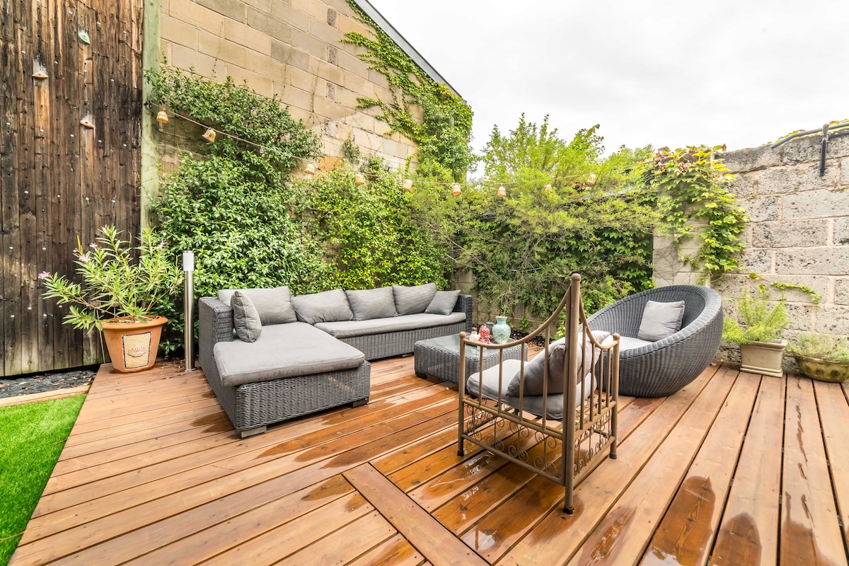 An outdoor seating area is presented on wooden decking, surrounded by lush greenery and stone walls. Comfortable gray seating is arranged with a round chair and a coffee table, creating an inviting space for relaxation. Soft lighting and potted plants enhance the tranquil environment.