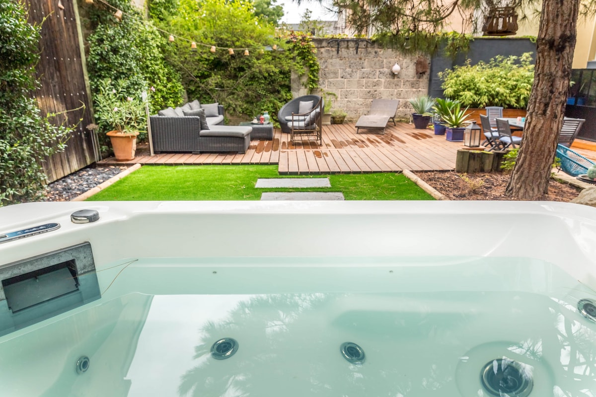 A private outdoor area features a jacuzzi surrounded by greenery, offering a serene retreat. Comfortable seating is arranged on a wooden deck, with lounge chairs spaced among potted plants. A well-maintained lawn adds to the inviting atmosphere.