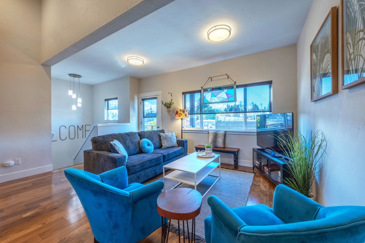 An inviting living area features a comfortable grey sofa accented by bright blue cushions. Two blue armchairs complement the space, while a light wooden coffee table sits in the center. Natural light enters through the windows, highlighting the warm wood flooring.