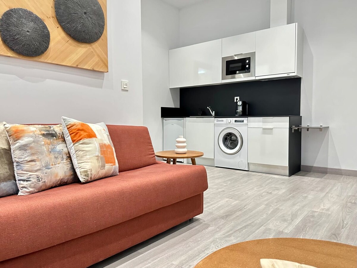 A modern suite apartment features a cozy seating area with a red sofa adorned with cushions. A compact kitchen with a microwave and washing machine is visible, complemented by a small round table. A neutral color palette enhances the overall design.
