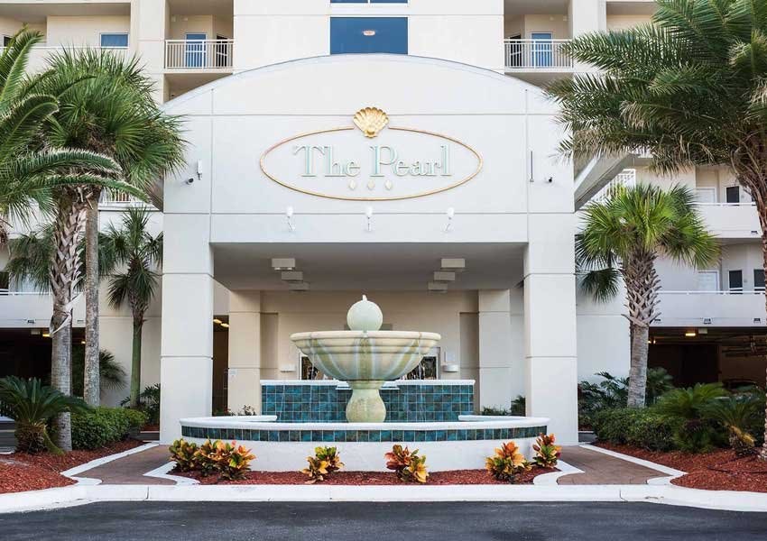 The entrance of The Pearl features a decorative fountain surrounded by lush green plants and colorful landscaping. A prominent sign displaying 'The Pearl' rests above the fountain, framed by the building's architecture that leads into the lobby area.