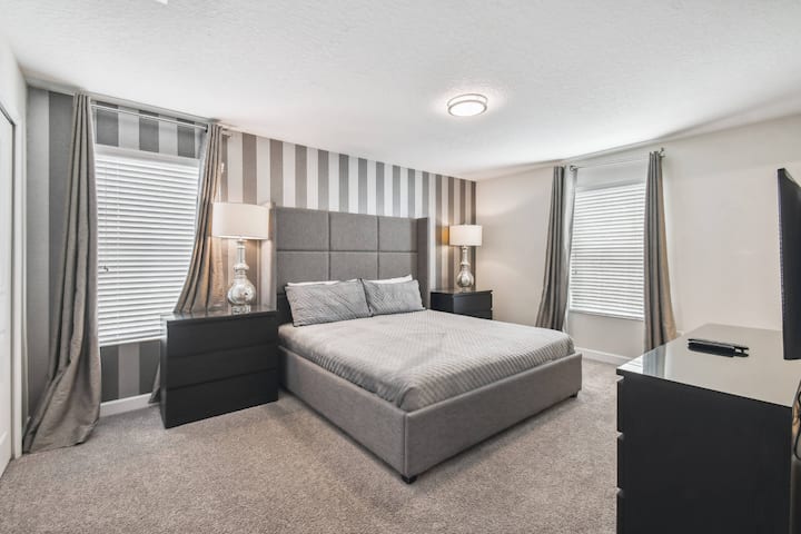 Zažite pokojný odpočinok s útulnou sivou posteľou obklopenou elegantnou pruhovanou tapetou a mäkkým prirodzeným svetlom. Elegantné nočné stolíky a moderné lampy umocňujú hrejivú a štýlovú atmosféru, ktorá je ideálna na relax a odpočinok.