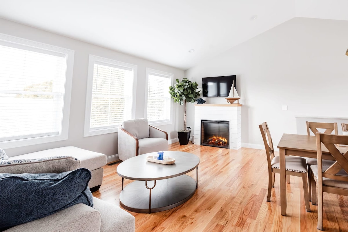 A bright living area features large windows allowing natural light to fill the space. A cozy seating arrangement includes two armchairs and a spacious sectional sofa. A circular coffee table sits in the center, accompanied by a dining table nearby. A decorative plant and an electric fireplace add warmth.
