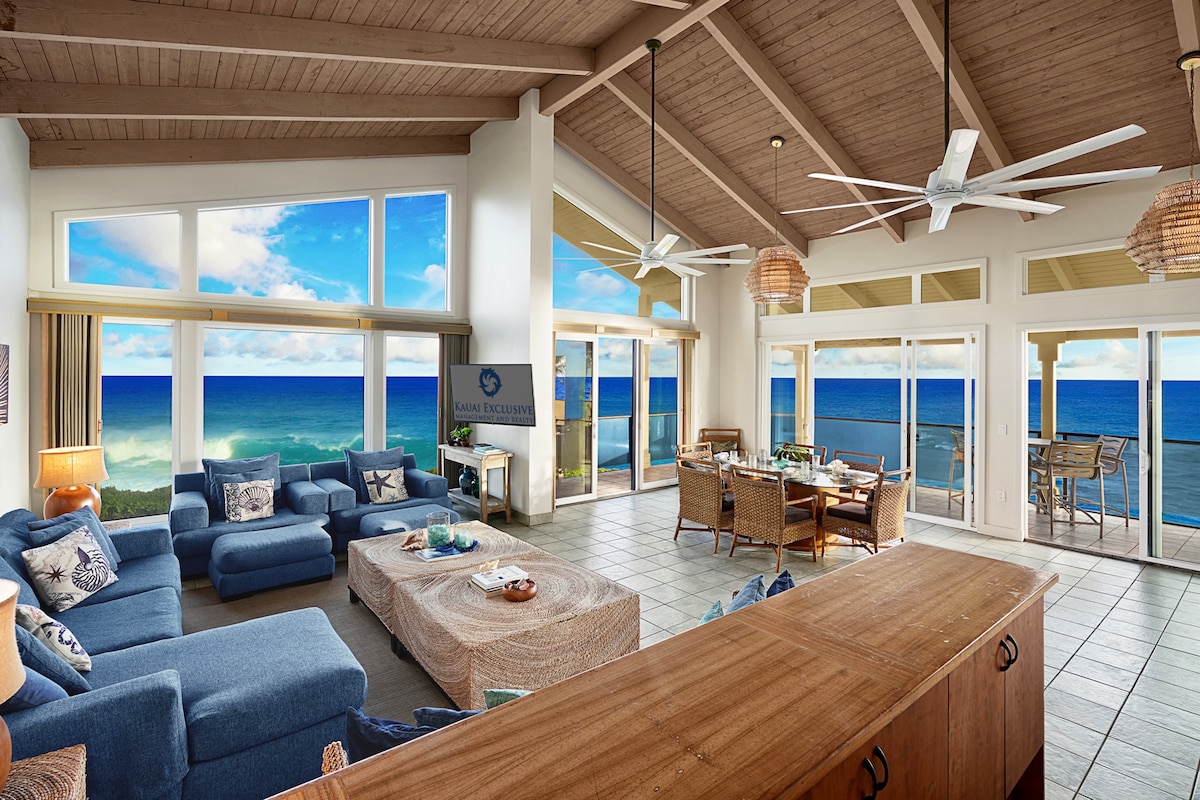 Expansive living area with high, wooden ceilings features large floor-to-ceiling windows that offer unobstructed ocean views. A comfortable sectional sofa is positioned around a coffee table. A dining area with a table and chairs is visible near sliding doors leading to the lanai.