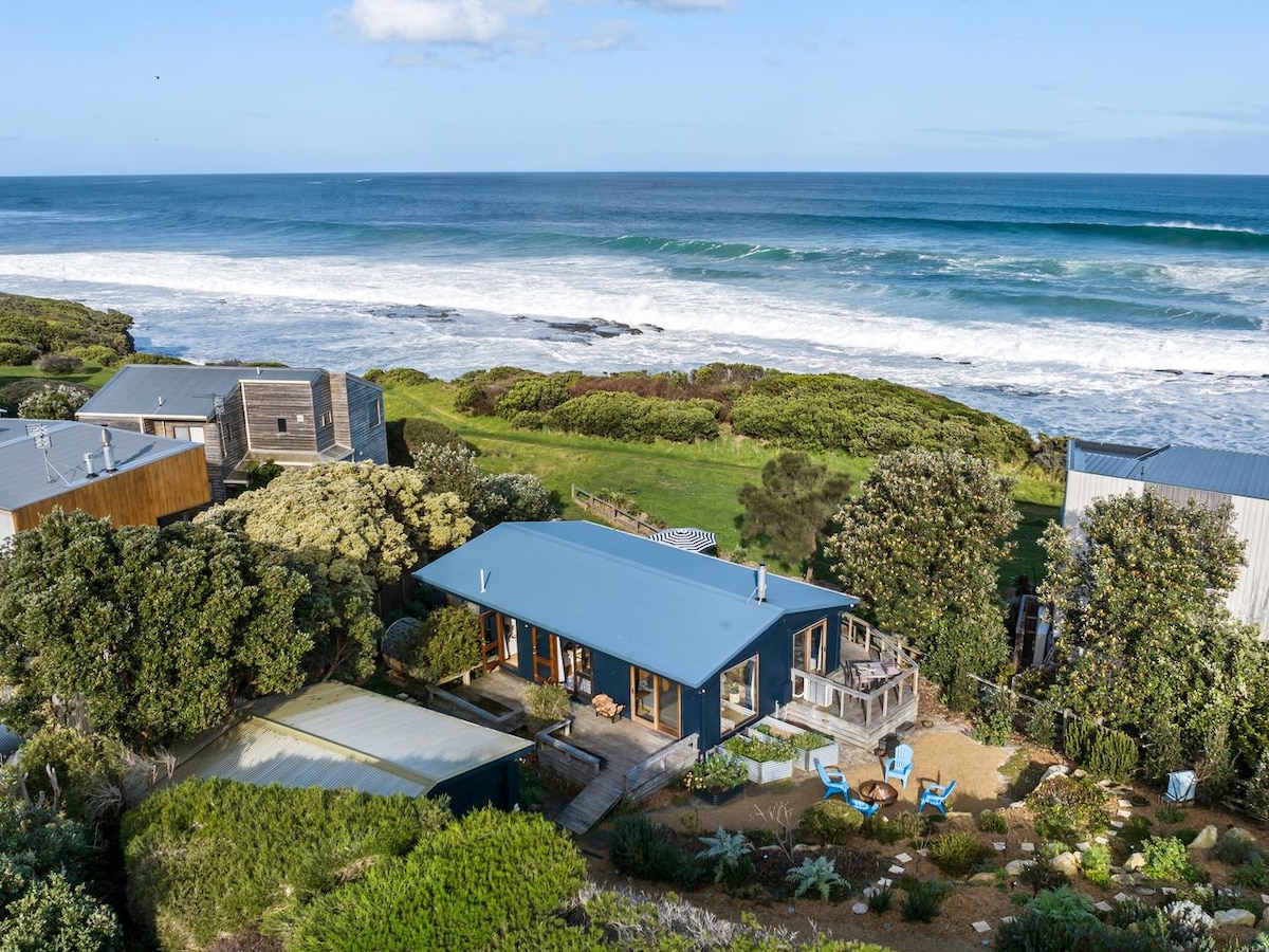 An aerial view captures a seaside retreat positioned close to the shoreline, surrounded by lush gardens and greenery. The calming ocean waves are visible in the background, while the cozy beach house features a blue roof, multiple decks, and inviting outdoor seating areas.