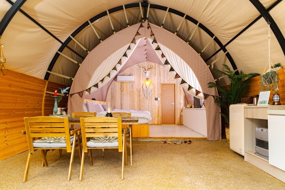 An inviting tent structure features a spacious interior with wooden accents and soft lighting. A cozy sleeping area is visible at the back, while a small dining space with wooden chairs and a table is set in the foreground, enhancing the warm atmosphere.