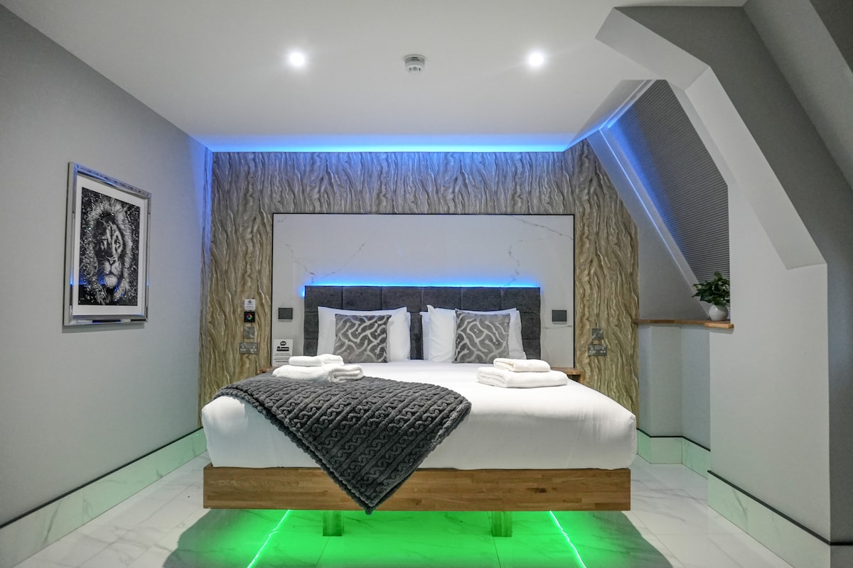 A modern bedroom is showcased with a spacious bed featuring a wooden frame and decorative grey throw. Subtle lighting accents the textured wall behind the bed. White marble flooring reflects the light, creating a serene environment, complemented by greenery and elegant artwork.